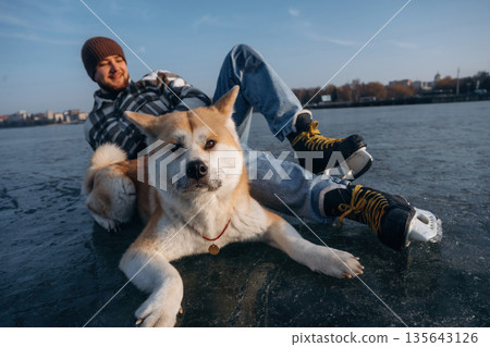 Just lying down and resting. Man with Akita Inu dog is on the ice with skates in winter Just lying down and resting. Man with Akita Inu dog is on the ice with skates in winter 135643126