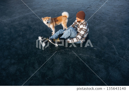 Be ready, preparing for the ride. Man with Akita Inu dog is on the ice with skates in winter Be ready, preparing for the ride. Man with Akita Inu dog is on the ice with skates in winter 135643140