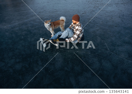 Be ready, preparing for the ride. Man with Akita Inu dog is on the ice with skates in winter Be ready, preparing for the ride. Man with Akita Inu dog is on the ice with skates in winter 135643141
