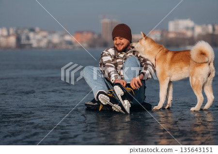 Ties shoelaces, sitting. Man with Akita Inu dog is on the ice with skates in winter 135643151