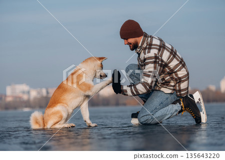 Learning the tricks, giving a paw. Man with Akita Inu dog is on the ice with skates in winter Learning the tricks, giving a paw. Man with Akita Inu dog is on the ice with skates in winter 135643220