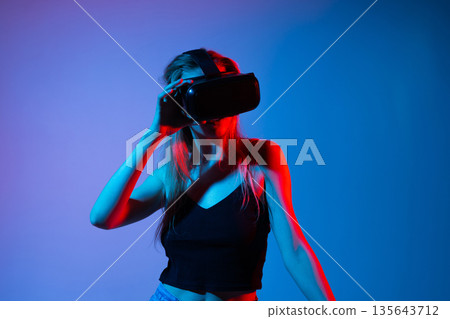 Woman touching virtual interface using VR headset, immersive virtual reality experience with futuristic blue and red neon lighting 135643712