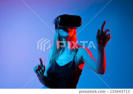 Woman Pointing Upward While Wearing Virtual Reality Headset, Futuristic VR Experience Concept With Neon Red and Blue Studio Lighting Woman Pointing Upward While Wearing Virtual Reality Headset, Futuristic VR Experience Concept With Neon Red and Blue Studio Lighting 135643713