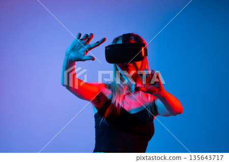 Wide shot of woman in VR headset interacting in virtual space, neon red blue light, immersive metaverse and gesture control 135643717
