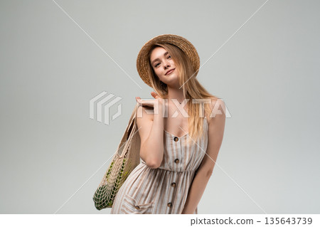 Stylish young woman posing with reusable grocery bag and straw hat, eco friendly summer lifestyle portrait on gray background 135643739