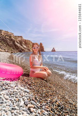 Beach woman vacation young woman in white swimsuit and sunglasses sitting on a pebble beach near the ocean enjoying summer holiday 135644681
