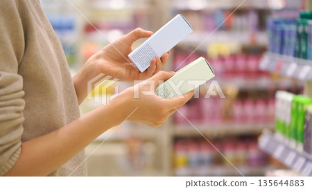 Customer Reading the Ingredient Labels Compares Two Skincare Products in Beauty Store. Close-up of a Female Hands, Woman Choosing Cosmetic Product. Makeup Selection. Conscious Choice. Mindful Shopping Customer Reading the Ingredient Labels Compares Two Skincare Products in Beauty Store. Close-up of a Female Hands, Woman Choosing Cosmetic Product. Makeup Selection. Conscious Choice. Mindful Shopping 135644883