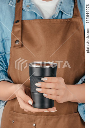 Woman holding reusable cup of hot drink wearing apron 135644978