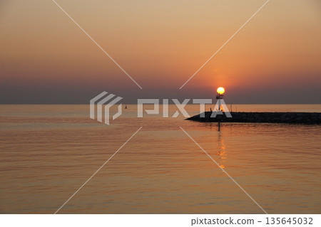 A huge ball of the sun, orange in color, rises from behind the horizon, against the backdrop of a stone pier.Dawn at sea. A huge ball of the sun, orange in color, rises from behind the horizon, against the backdrop of a stone pier.Dawn at sea. 135645032