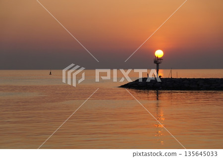 Dawn at sea.A huge ball of the sun, orange in color, rises from behind the horizon, against the backdrop of a stone pier  135645033