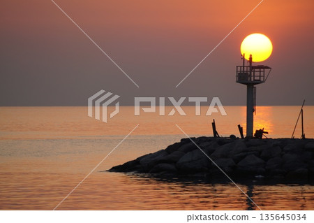 A huge ball of the sun, orange in color, rises from behind the horizon, against the backdrop of a stone pier and a lighthouse.Dawn at sea. 135645034