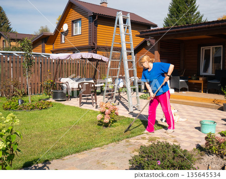 Woman raking cut grass on paved patio near green lawn and country house. Autumn horticulture work in backyard. Outdoor house chore concept. Woman raking cut grass on paved patio near green lawn and country house. Autumn horticulture work in backyard. Outdoor house chore concept. 135645534
