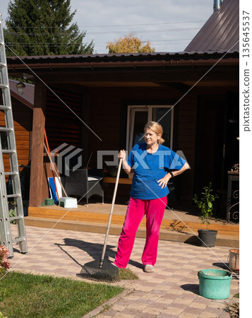 Woman stands with rake on paved patio beside house, ready for autumn garden cleanup work Woman stands with rake on paved patio beside house, ready for autumn garden cleanup work 135645537