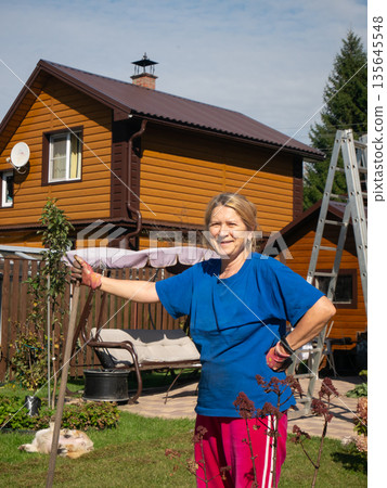 Woman gardener in blue t-shirt and pink pants smiling in backyard garden. Happy female homeowner working on landscaping project. 135645548