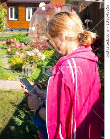 Woman using smartphone in her garden, browsing internet or checking social media, digital connection outdoors concept. Woman using smartphone in her garden, browsing internet or checking social media, digital connection outdoors concept. 135645658