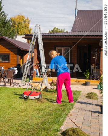 Woman mowing lawn with a red lawnmower in her backyard on a sunny day. Housekeeping and garden care concept. Outdoor activity. Woman mowing lawn with a red lawnmower in her backyard on a sunny day. Housekeeping and garden care concept. Outdoor activity. 135645659