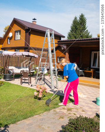 Woman raking cut grass in a garden with a large ladder near a house. Autumn cleanup concept of backyard maintenance and preparing for winter. 135645666