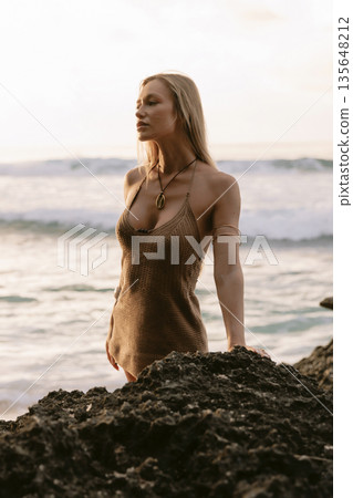 Blonde woman in knit dress standing on rocks by the ocean at sunset Blonde woman in knit dress standing on rocks by the ocean at sunset 135648212