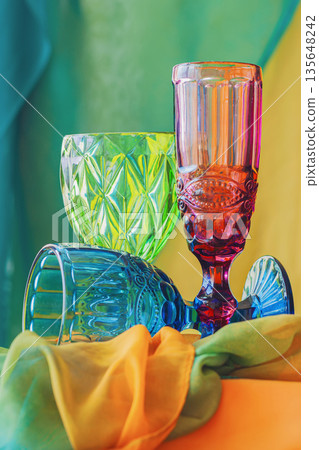 Colorful Glasses Still Life. Vibrant composition of multicolored vintage glassware on bright textile background. Concept of retro aesthetics, glass texture, colorful art lifestyle still life Colorful Glasses Still Life. Vibrant composition of multicolored vintage glassware on bright textile background. Concept of retro aesthetics, glass texture, colorful art lifestyle still life 135648242