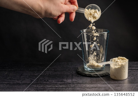 Protein drink. Hand pouring protein powder in a drinking glass. Dark background. Body building, muscle growth and food supplement concept Protein drink. Hand pouring protein powder in a drinking glass. Dark background. Body building, muscle growth and food supplement concept 135648552