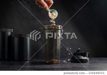 Protein drink. Hand pouring protein powder in a black shaker. Plastic tube on dark background. Body building, muscle growth and food supplement concept Protein drink. Hand pouring protein powder in a black shaker. Plastic tube on dark background. Body building, muscle growth and food supplement concept 135648553