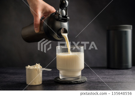 Protein drink. Pouring protein shake in a glass. Dark background. Body building, muscle growth and food supplement concept Protein drink. Pouring protein shake in a glass. Dark background. Body building, muscle growth and food supplement concept 135648556
