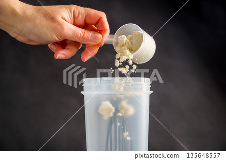 Protein drink. Hand pouring protein powder in a white shaker. Dark background. Body building, muscle growth and food supplement concept 135648557