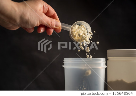 Protein drink. Hand pouring protein powder in a white shaker. Plastic tube on dark background. Body building, muscle growth and food supplement concept Protein drink. Hand pouring protein powder in a white shaker. Plastic tube on dark background. Body building, muscle growth and food supplement concept 135648564