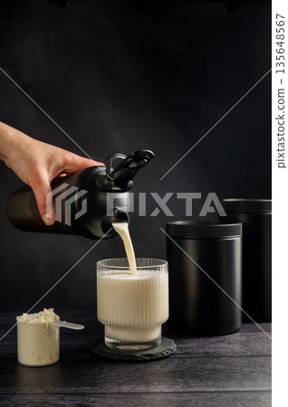 Protein drink. Pouring protein shake in a glass. Dark background. Body building, muscle growth and food supplement concept Protein drink. Pouring protein shake in a glass. Dark background. Body building, muscle growth and food supplement concept 135648567