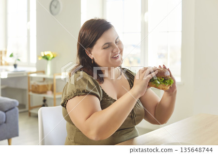 Smiling overweight woman enjoying delicious burger at home, indulging in fast food in kitchen. 135648744