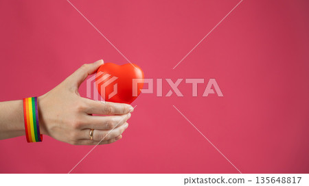 Woman with engagement ring and rainbow bracelet holding heart on pink background. Copy space. Woman with engagement ring and rainbow bracelet holding heart on pink background. Copy space. 135648817
