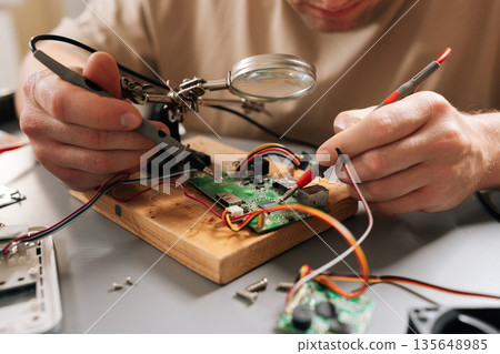 Close-up view of technician focusing on intricate details of circuit board on wooden base with magnifying glass, using helping hands tools and wires during electronic repair or assembly process. Close-up view of technician focusing on intricate details of circuit board on wooden base with magnifying glass, using helping hands tools and wires during electronic repair or assembly process. 135648985
