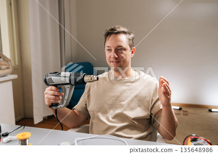 Young adult repairman holding heat gun, finding happy in diy repairs and electronics work at home, feeling excited about new project in workshop space. Concept of modern technology. Young adult repairman holding heat gun, finding happy in diy repairs and electronics work at home, feeling excited about new project in workshop space. Concept of modern technology. 135648986
