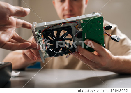 Close-up of repairman completing electronic repairs in bright workshop, holding disassembled graphic card, exposing intricate internal components and ensuring proper functionality of hardware. Close-up of repairman completing electronic repairs in bright workshop, holding disassembled graphic card, exposing intricate internal components and ensuring proper functionality of hardware. 135648992
