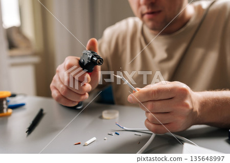 Man heats stripped electrical cable with gas torch while soldering and repairing electronics at cluttered workbench, focused on precise diy wiring and connection work. 135648997