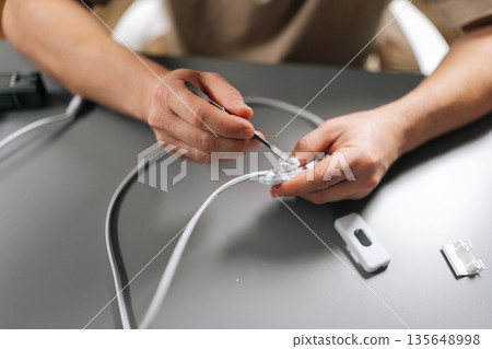 Hands using small tool to open white electrical switch on cable, showcasing process of repairing home electronics and demonstrating electrical work. Concept of hardware, repairing, technology. Hands using small tool to open white electrical switch on cable, showcasing process of repairing home electronics and demonstrating electrical work. Concept of hardware, repairing, technology. 135648998