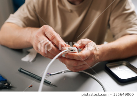 Close-up of male hands connecting white electrical cable to small housing unit, highlighting process of repairing and maintaining electronics, home wiring, technology infrastructure. 135649000