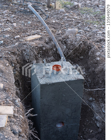 Concrete foundation block with anchor bolts and orange PVC pipe in a trench. Construction site infrastructure detail Concrete foundation block with anchor bolts and orange PVC pipe in a trench. Construction site infrastructure detail 135649079
