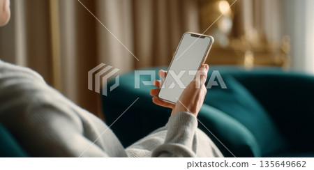 Person holding smartphone with blank screen while sitting on teal sofa in cozy indoor setting with blurred background 135649662