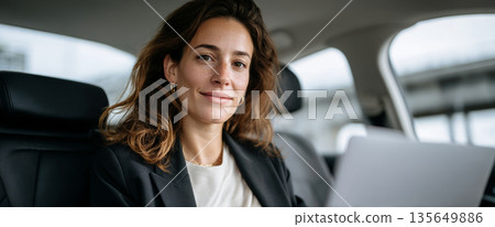 Confident young woman sitting in car backseat working on laptop with relaxed expression and casual business attire 135649886