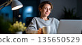 Smiling woman working on laptop at home in cozy sweater holding coffee cup with desk lamp and blurred background at night 135650529
