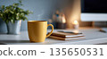 Steaming yellow coffee mug on white desk with blurred background of notebooks, computer, candle, and plant in cozy workspace setting 135650531