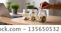 Hand placing coin into glass jar with coins on wooden table near laptop and blurred plants in background 135650532