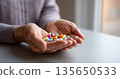 Close-up of elderly hands holding various colorful pills and capsules indoors near window with soft natural light 135650533