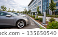 Electric cars parked and charging at modern outdoor charging stations near building with greenery and cloudy sky 135650540