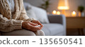 Close-up of elderly person holding white pills in hands sitting on couch in cozy living room with warm lighting 135650541