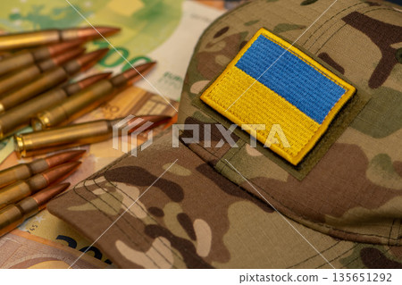 Military cap with Ukraine flag placed near ammunition and euro banknotes. 135651292