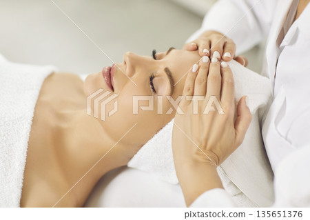 Close up photo of pretty young woman lying with closed eyes and having face or head massage in spa 135651376