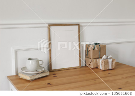 Minimal interior mockup with blank vertical wooden picture frame on table. Elegant home decor scene with coffee cup, books and gift boxes. Neutral Scandinavian style workspace, birthday lifestyle view 135651608