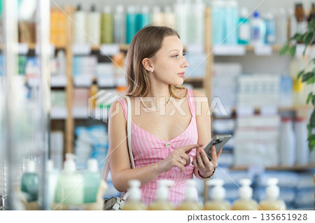 Female shopper searches for necessary goods on the shelves of a supermarket, checking list on her smartphone Female shopper searches for necessary goods on the shelves of a supermarket, checking list on her smartphone 135651928